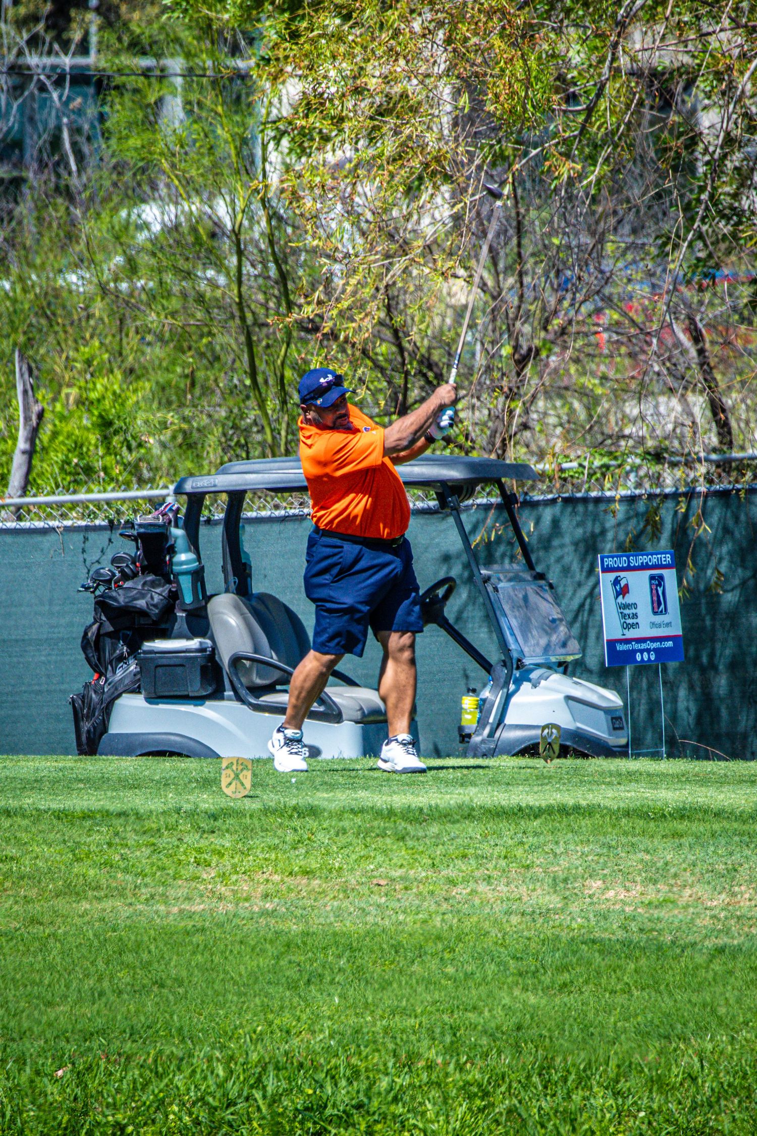 Golfer in orange shirt and blue shorts swings club on green course with golf cart in background.
