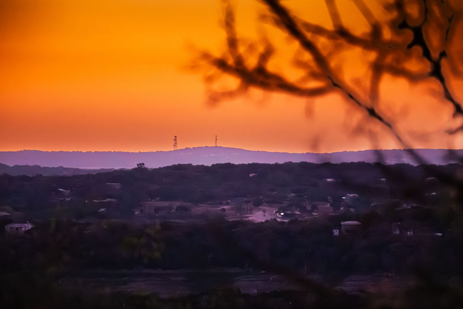 Orange sunset over distant mountains with silhouetted tree branches in the foreground.