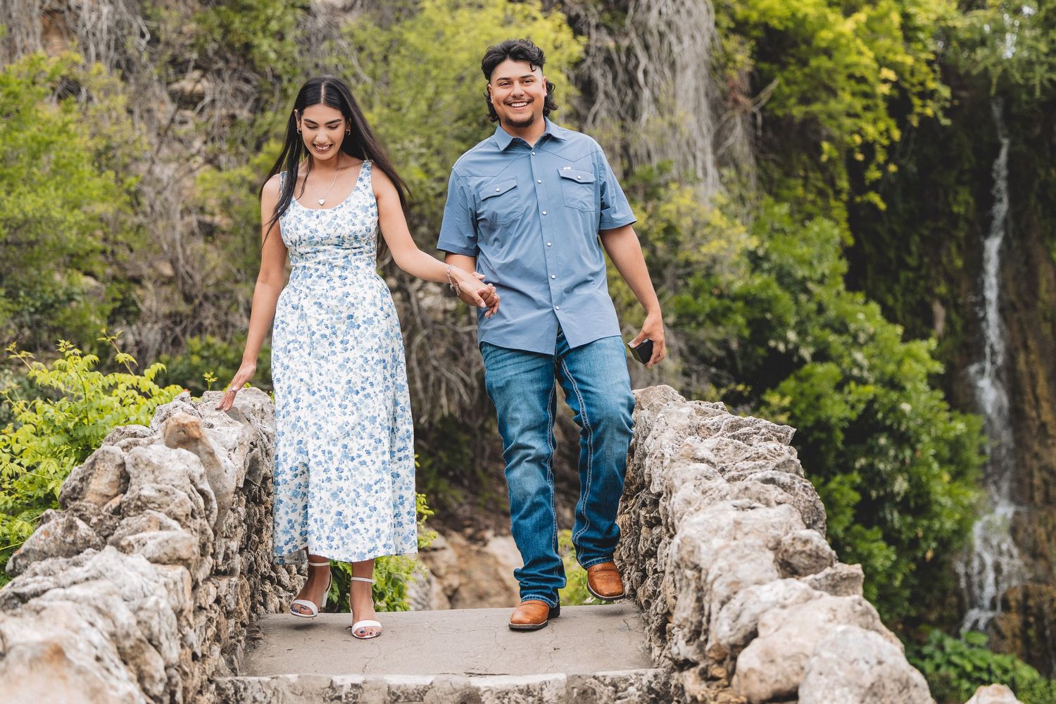 Couple holding hands walking down stone steps with a waterfall and lush greenery in the background.