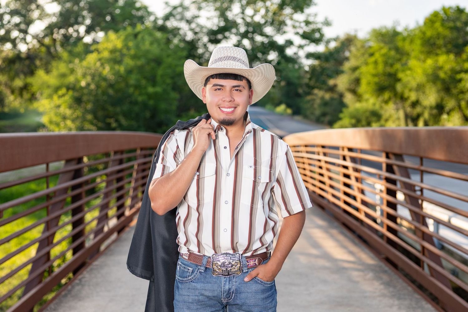 Young man in cowboy hat and striped shirt smiling on a bridge, holding a jacket outdoors.