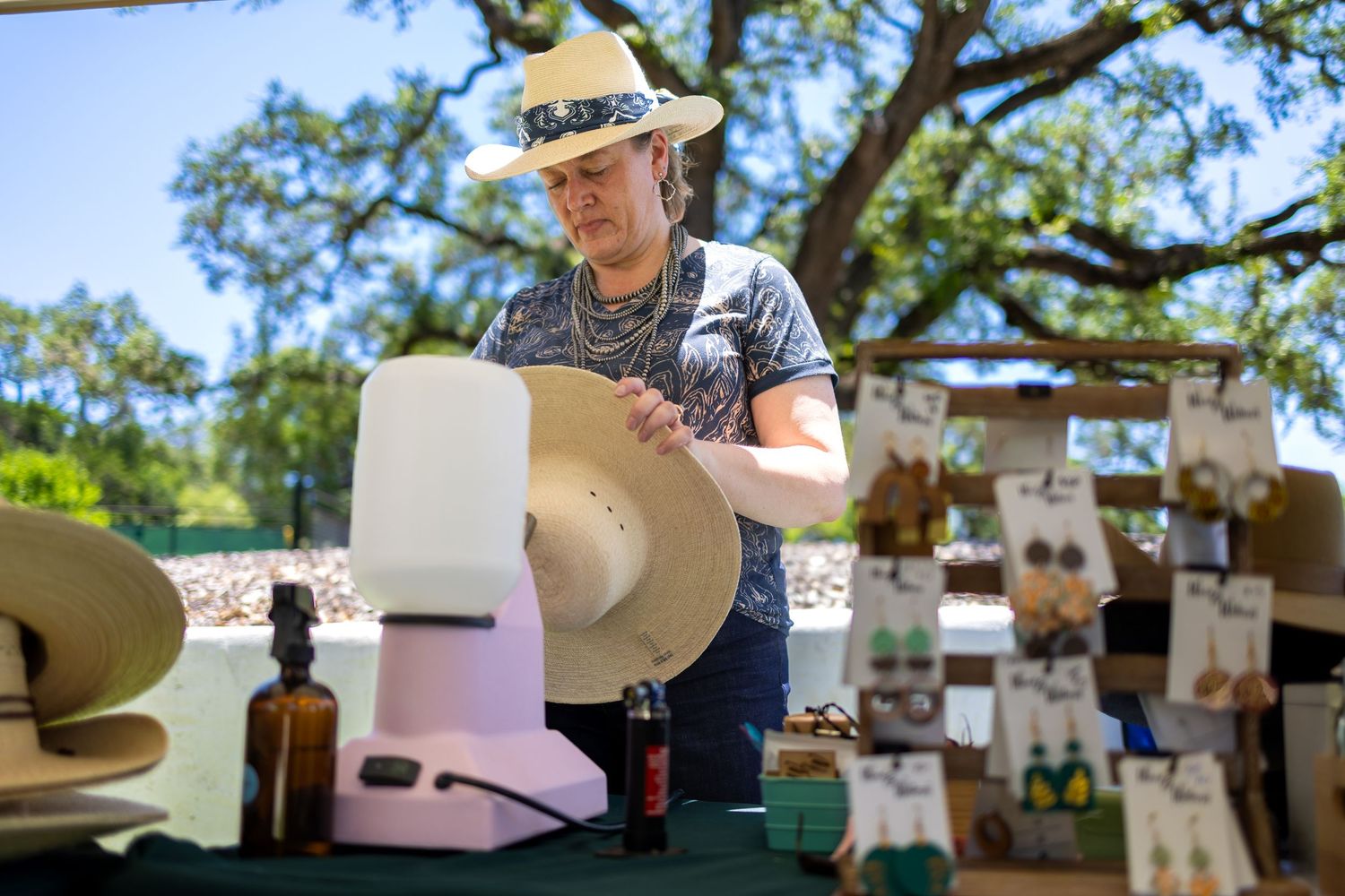 Artisan customizing a straw hat at outdoor market booth with accessories on display.