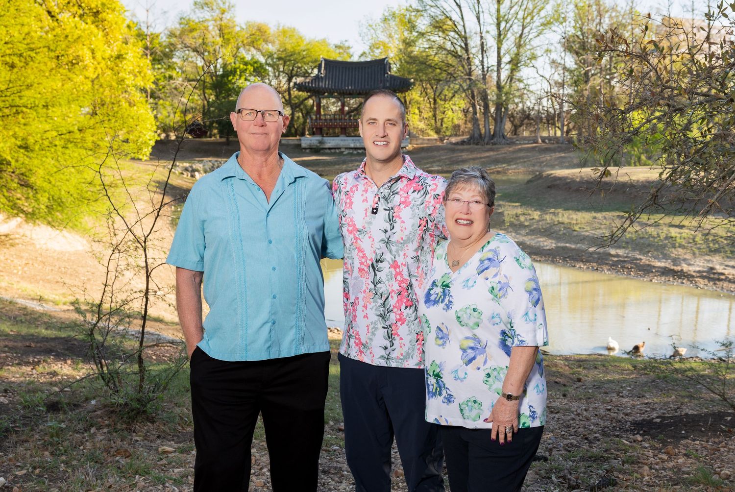 A family of three standing in a park with trees and a body of water in the background.