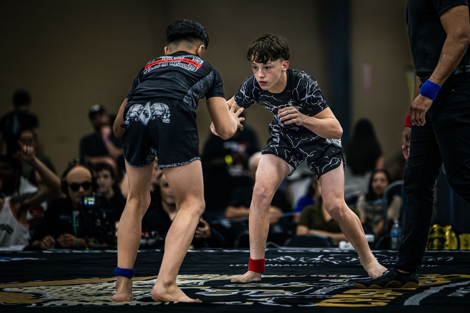 Two young MMA fighters compete in a grappling match at an indoor martial arts tournament.