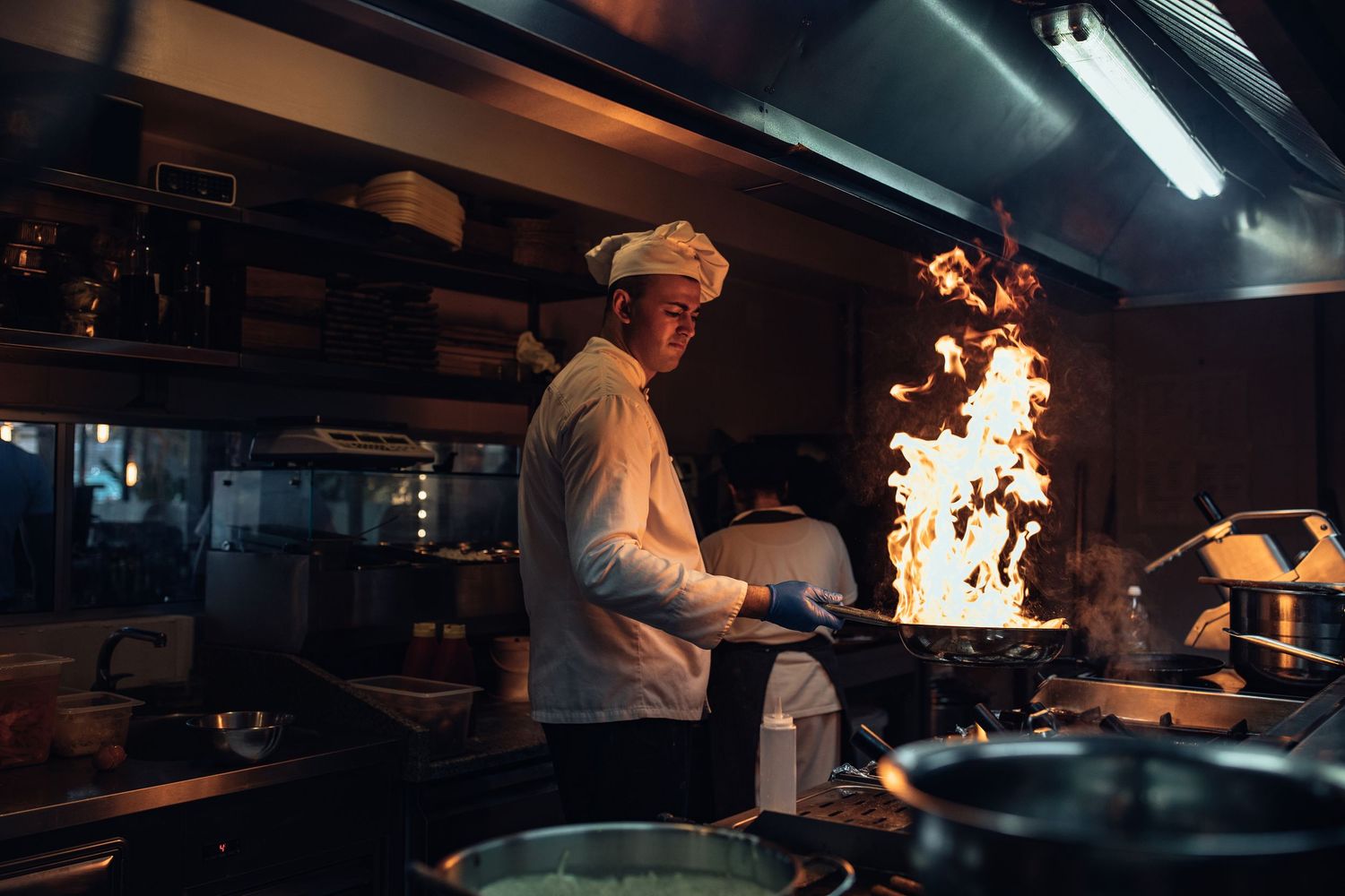Chef in white uniform cooking with dramatic flames in a professional restaurant kitchen.