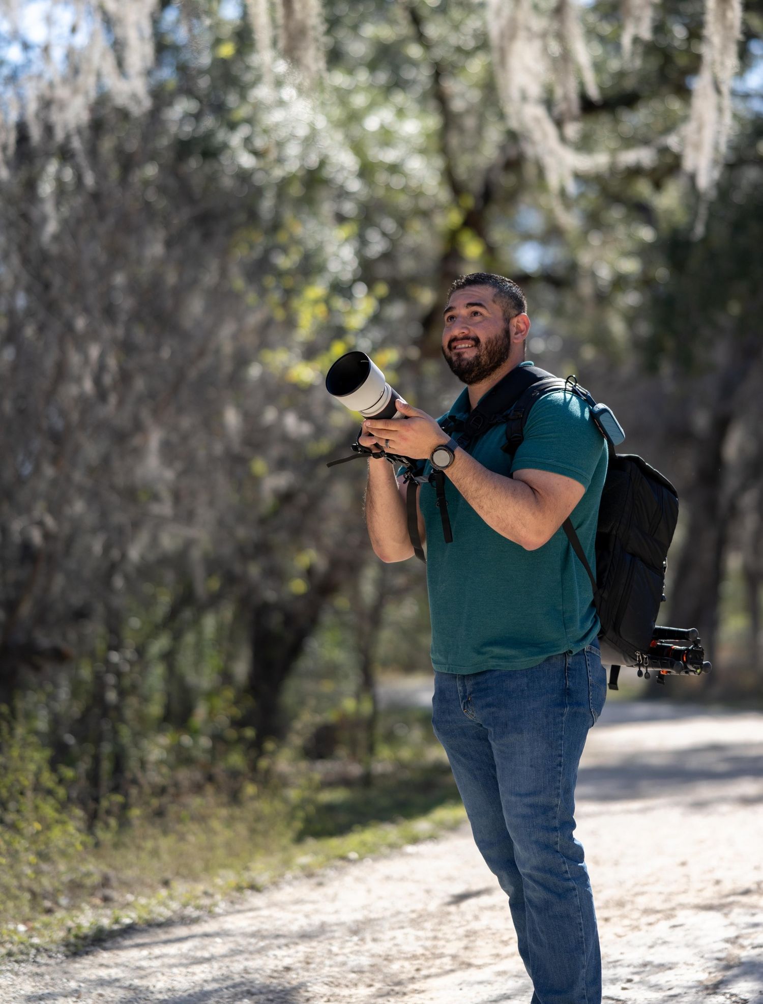 Man with camera and large telephoto lens walking on a forest path with Spanish moss hanging from trees.