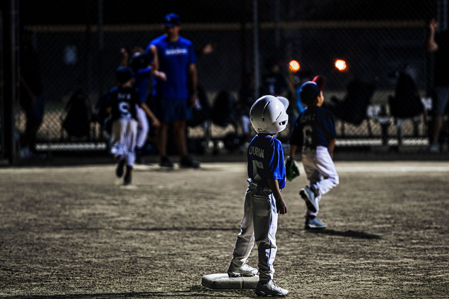 Young children in baseball uniforms play a night game on a lit field, with a small player standing on base in the foreground.