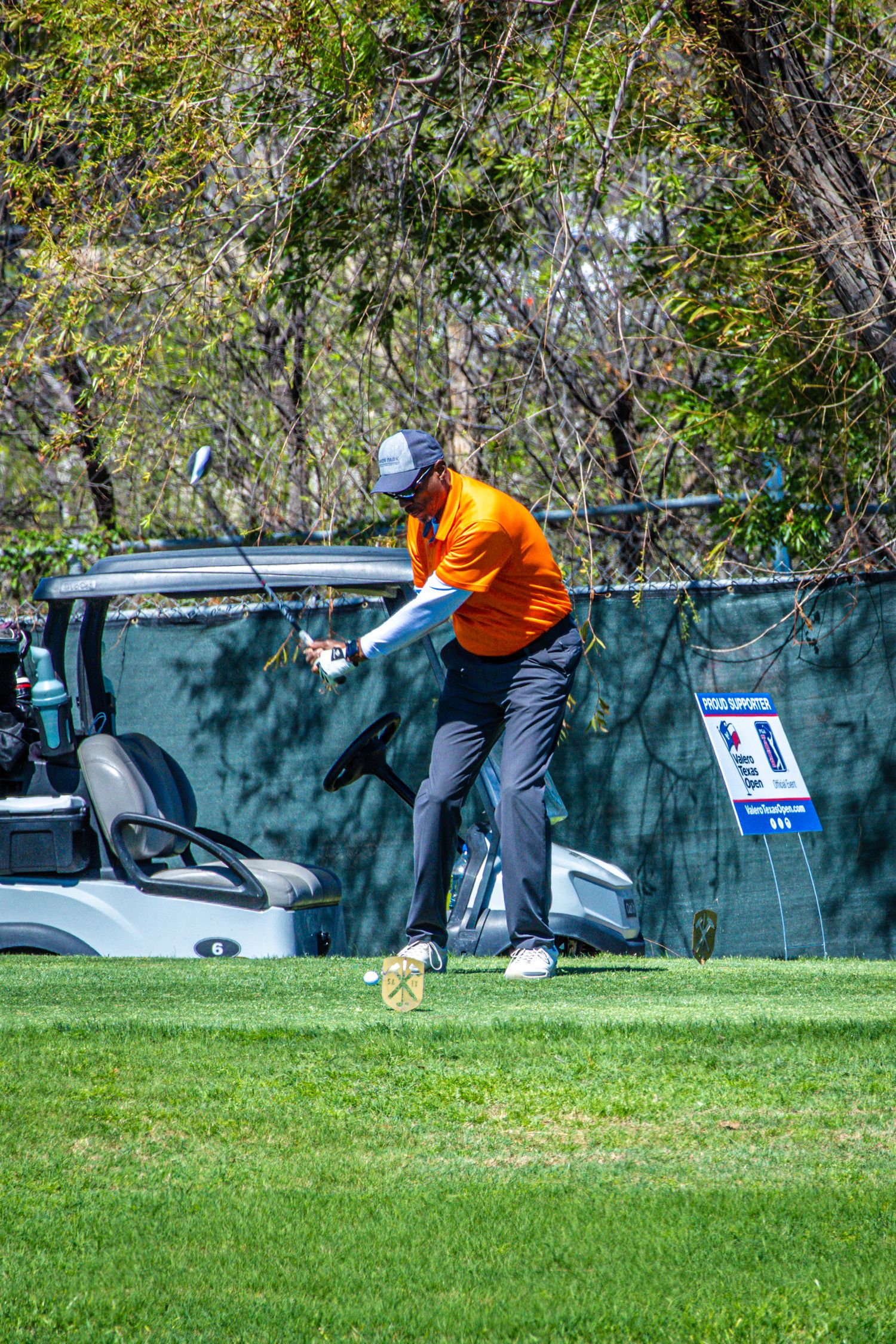 Golfer in orange shirt swinging a club on a green course with a golf cart in the background.