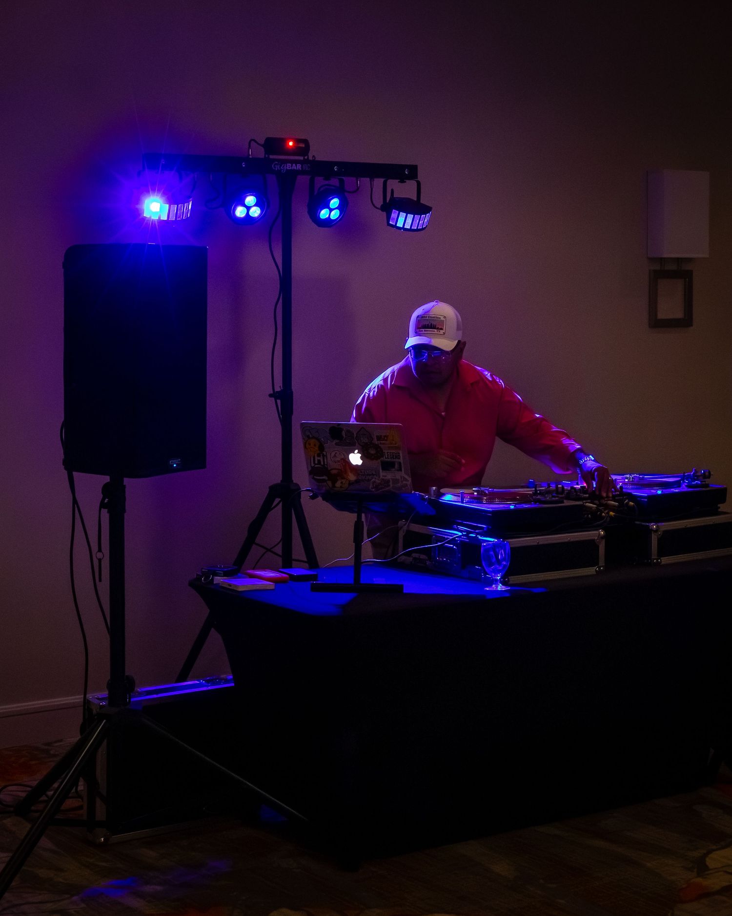 DJ performing at an event with blue stage lighting, turntables, laptop, and speaker on a tripod in a dimly lit room.