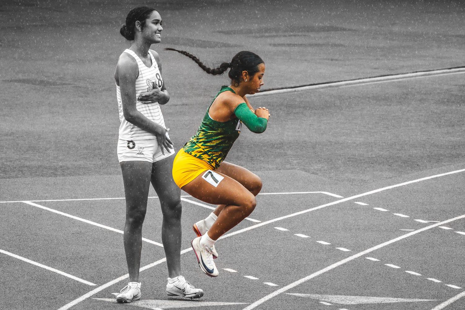 Female sprinter in yellow-green uniform runs on track while another athlete stands in black and white background.