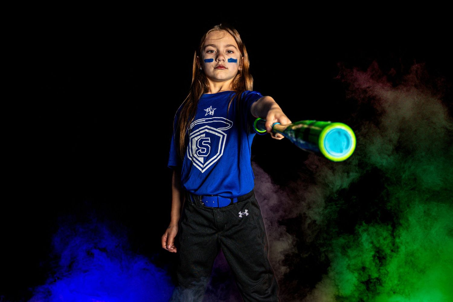Young girl in blue softball uniform holds bat amid colorful smoke clouds in dramatic sports portrait.