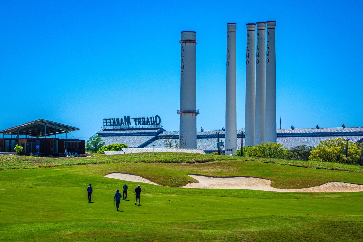 Golfers on a scenic golf course with sand bunkers near an industrial building with tall chimneys.