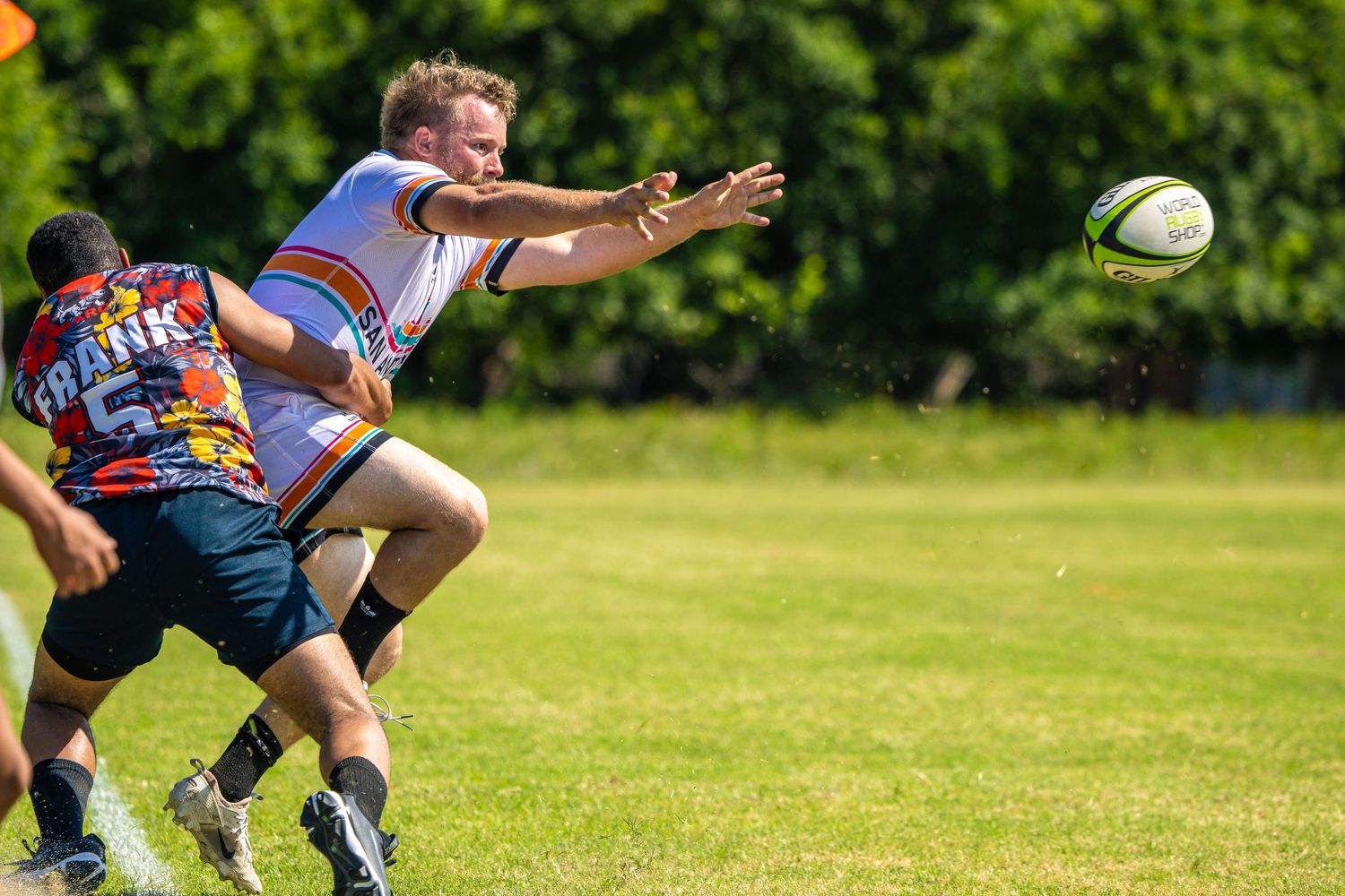 Rugby player passing ball mid-game on green field during sunny outdoor match with teammates in colorful jerseys.