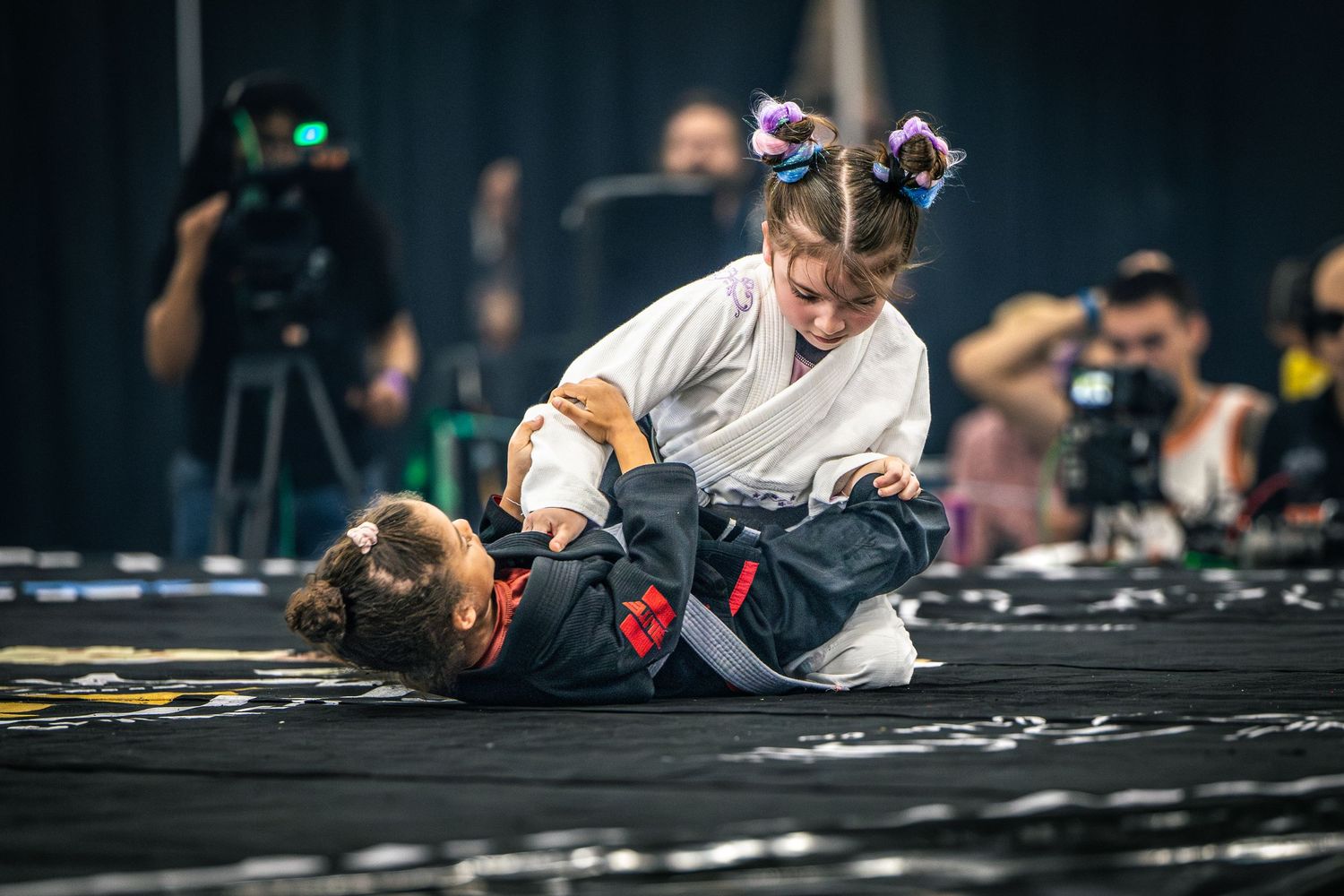 Two children competing in a Brazilian jiu-jitsu match on a black mat at a martial arts tournament.