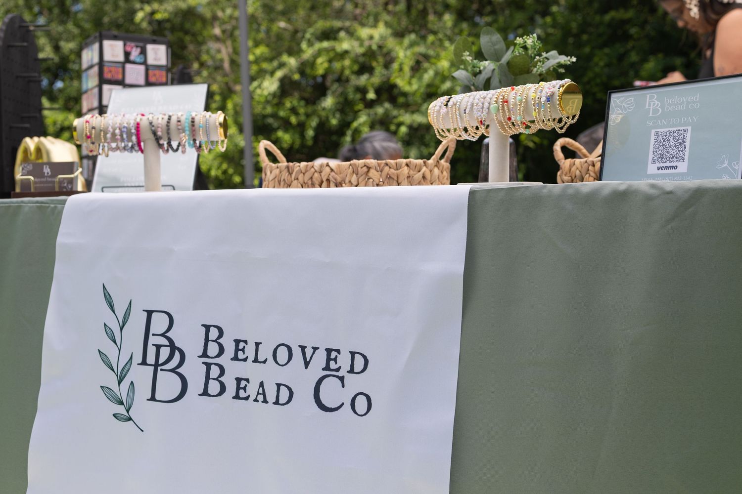 Beloved Bead Co vendor booth display with jewelry and accessories on a green draped table at an outdoor market.