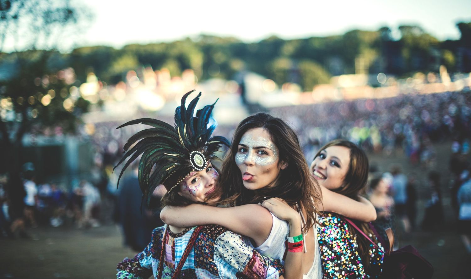 Three festival-goers in colorful outfits and feathered headpiece embrace and smile at an outdoor music festival.