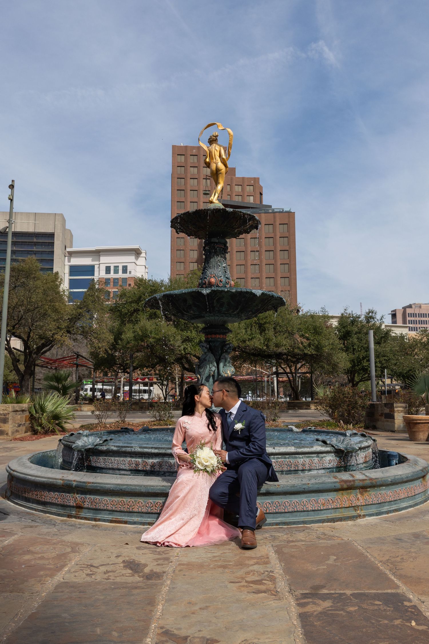 A couple poses for romantic photos at a circular fountain plaza with city buildings and blue sky in the background.