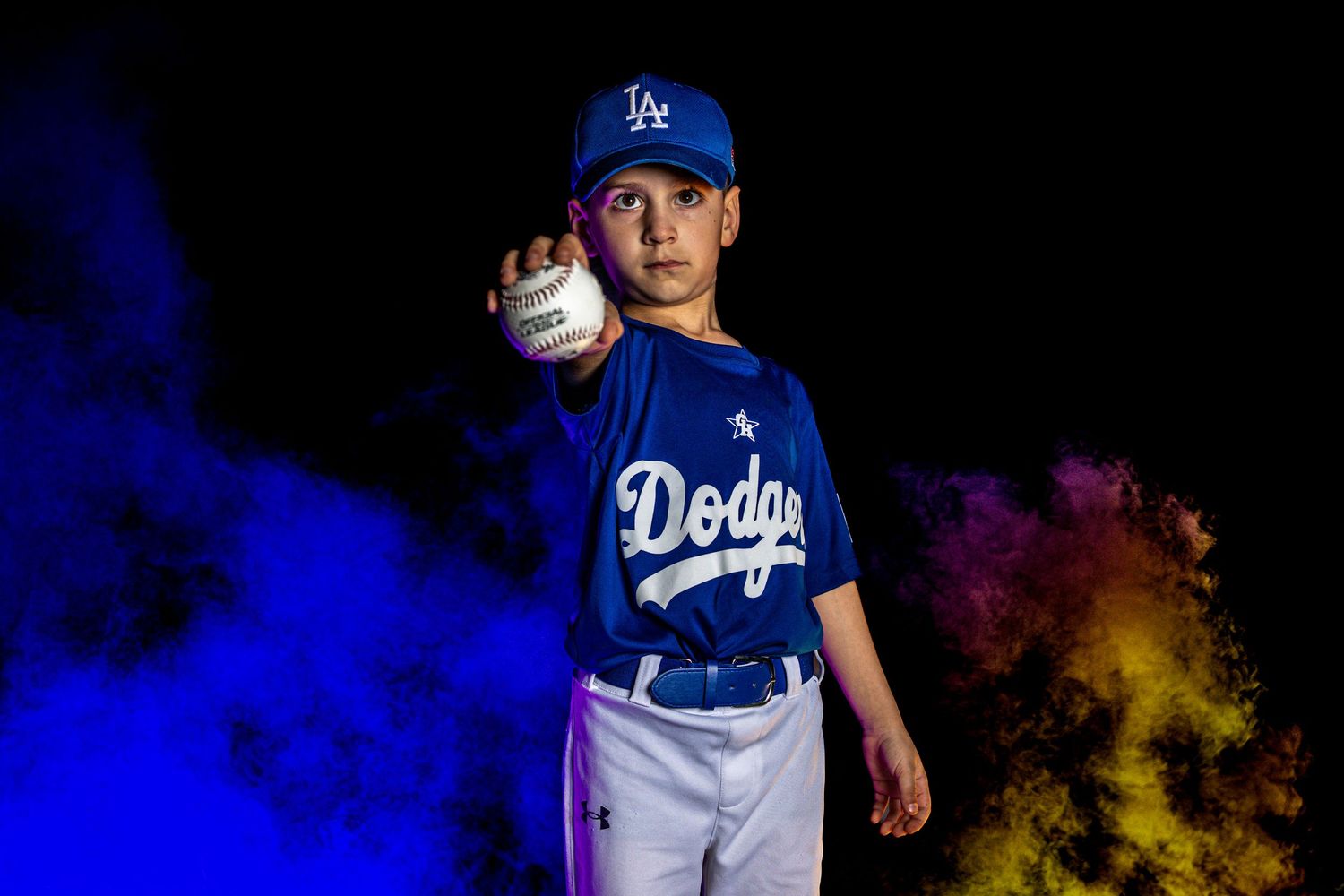 Young boy in LA Dodgers baseball uniform poses dramatically holding a ball with colorful smoke behind him.