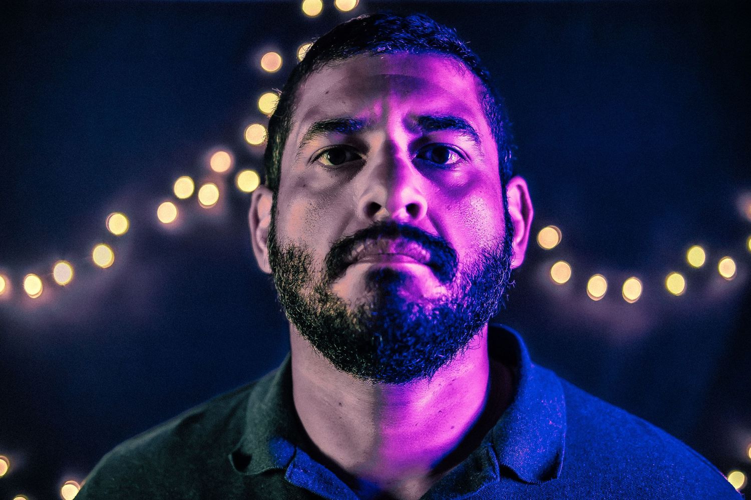 Bearded man illuminated by purple light with bokeh string lights glowing in dark background.