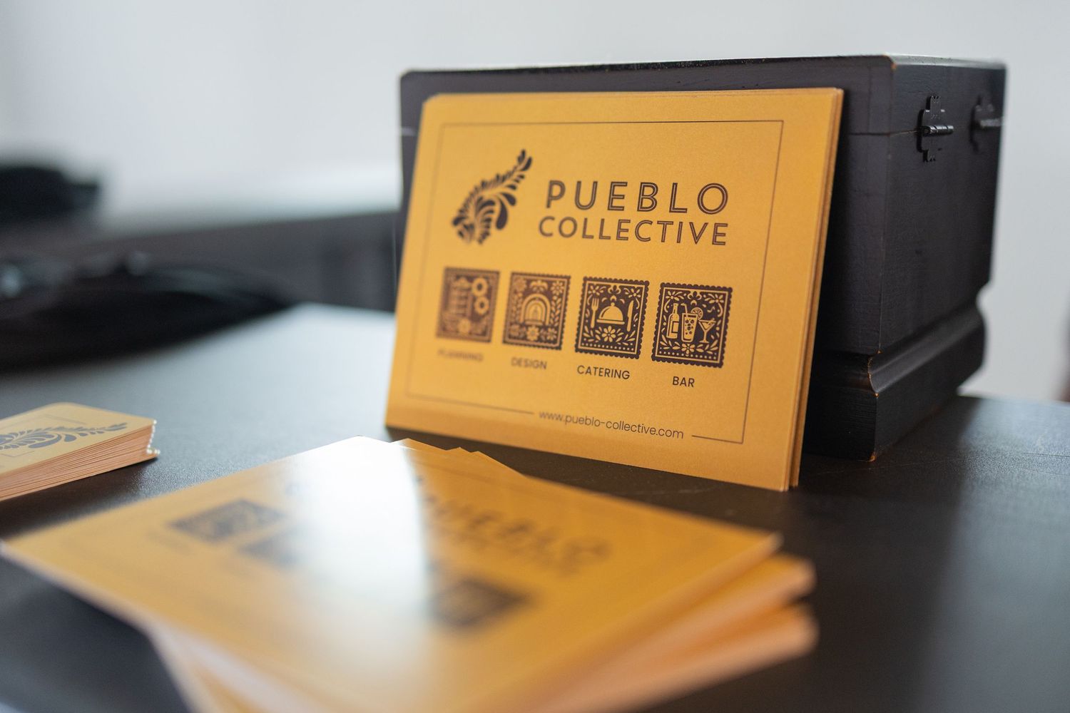 Pueblo Collective branded business cards and black box displayed on a wooden desk.