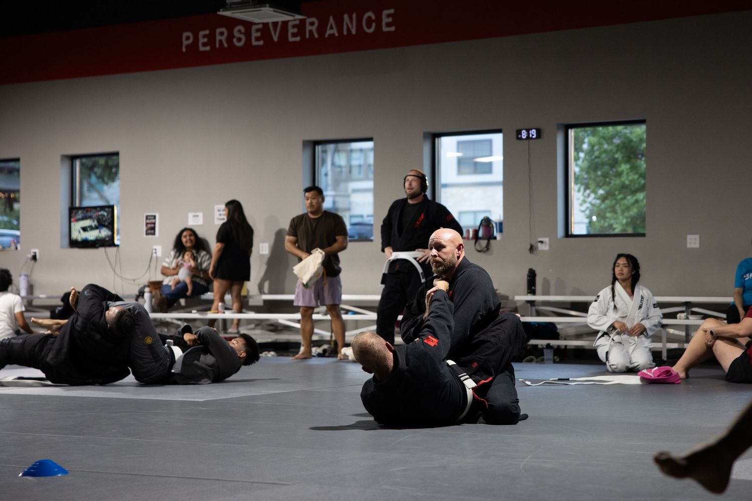 Martial arts students practice BJJ grappling techniques on a mat in a gym with Perseverance sign on wall.