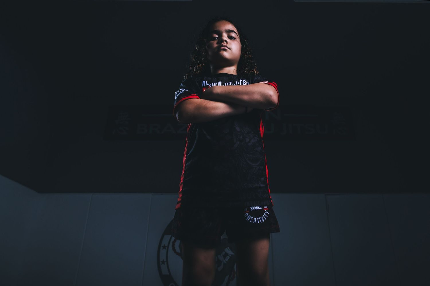 Female MMA fighter poses confidently with arms crossed wearing black and red fight shorts in dark gym.