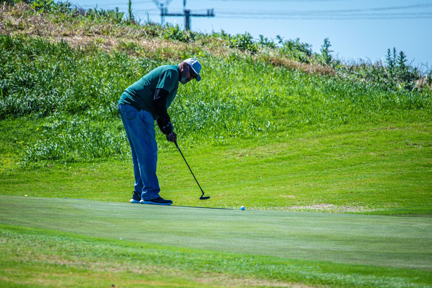 A golfer in a green shirt and blue pants putts on a lush green golf course on a sunny day.