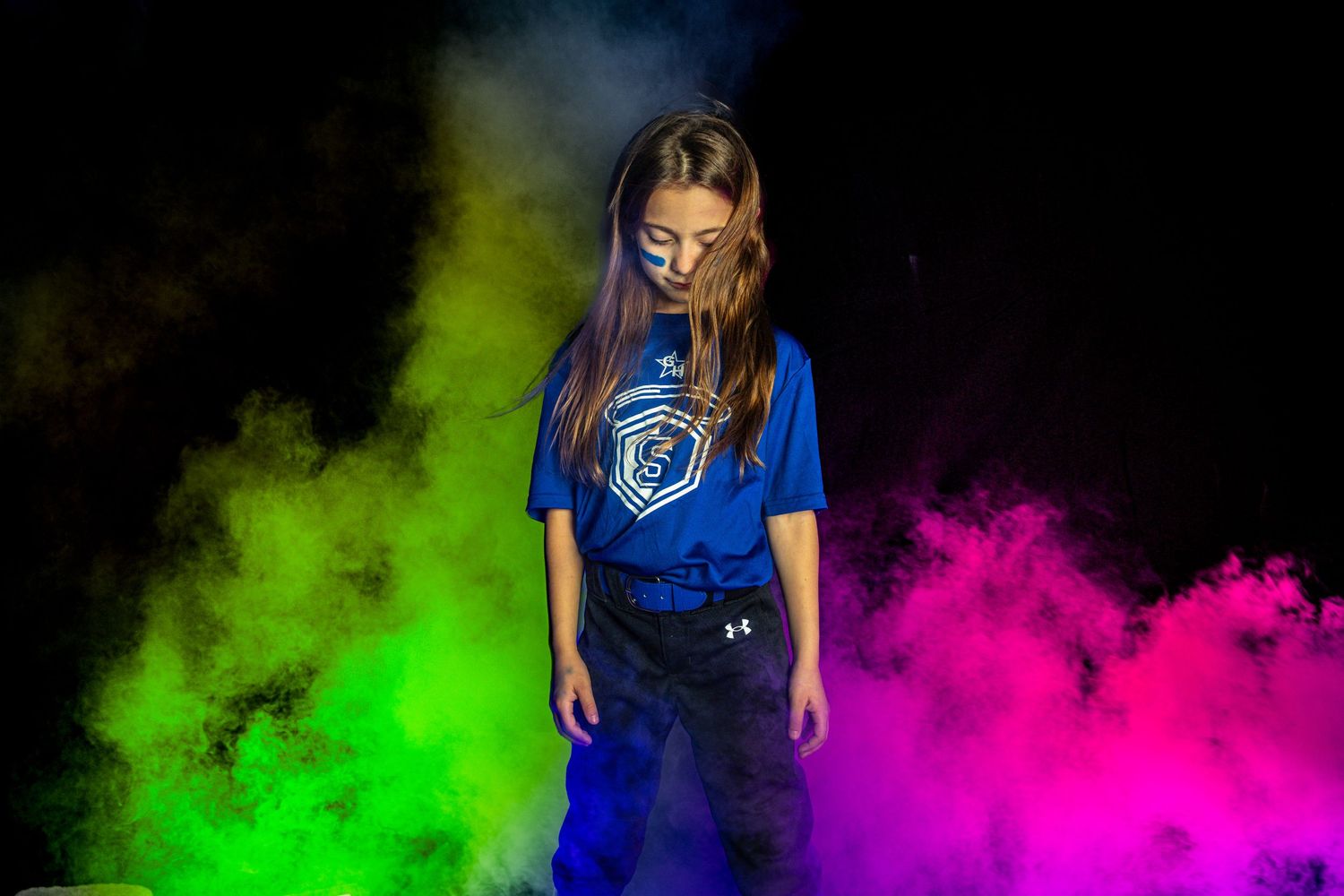 Young girl in blue softball jersey surrounded by vibrant green, pink, and yellow color powder clouds on dark background.