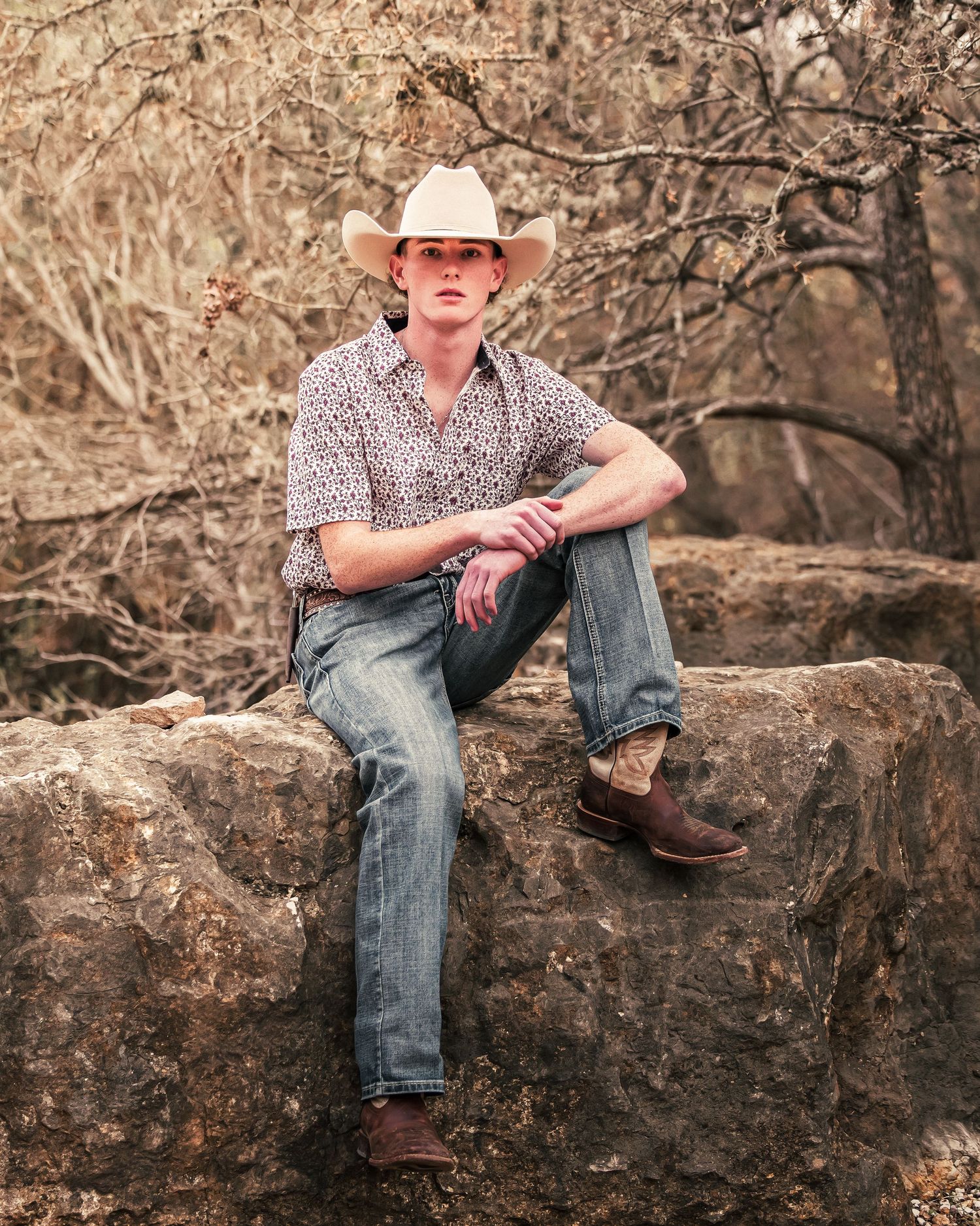 Person in white hat and jeans sitting casually on large rocks in a rustic outdoor setting with dried branches in background.