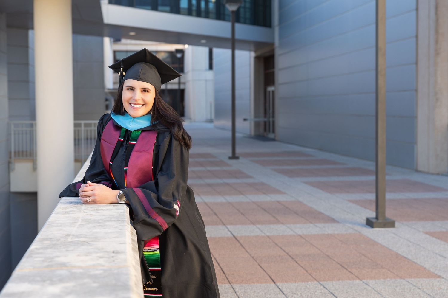 Graduate in cap and gown poses with diploma outside modern building on graduation day.
