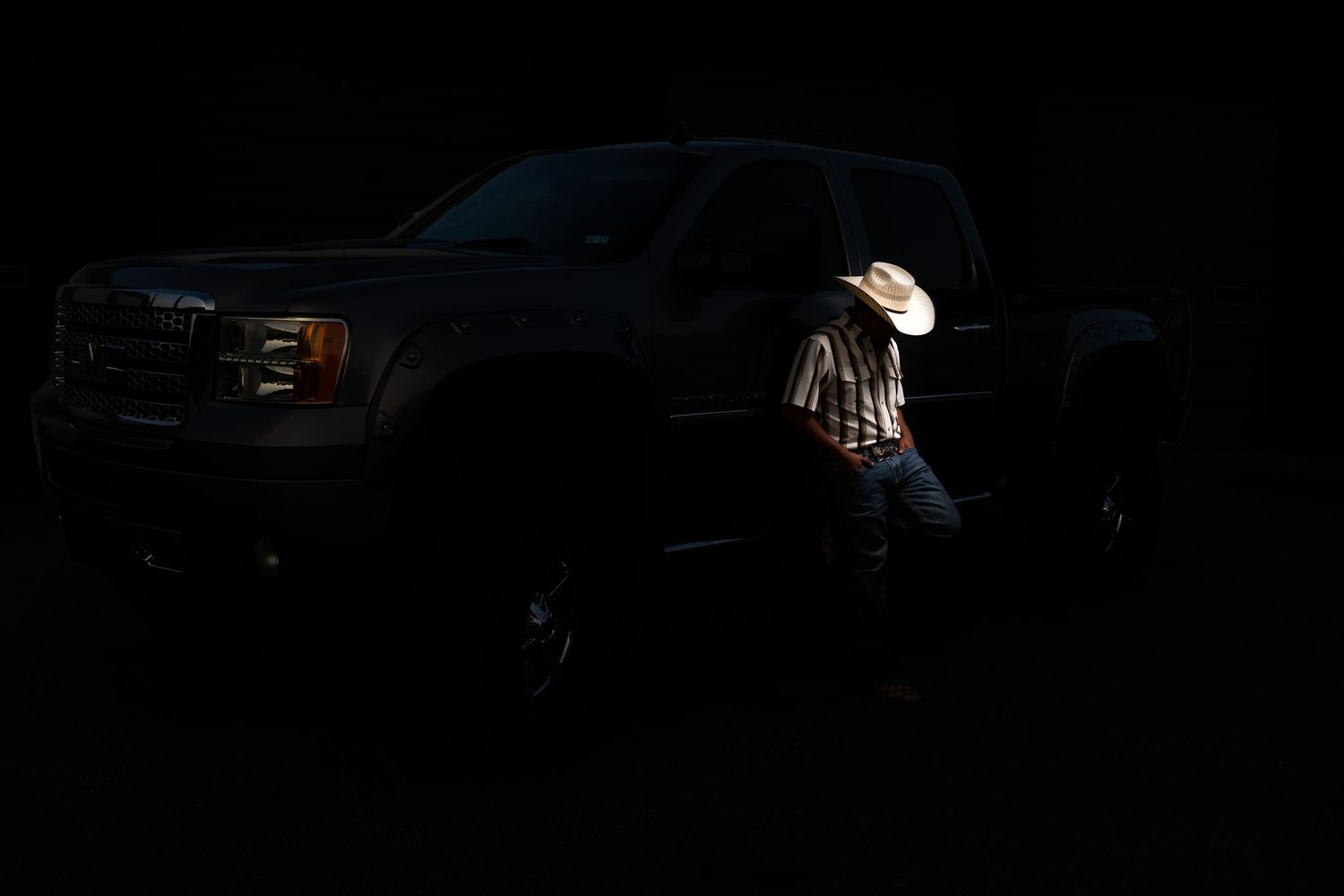 A cowboy in a white hat leans against a truck in dramatic low-key lighting.