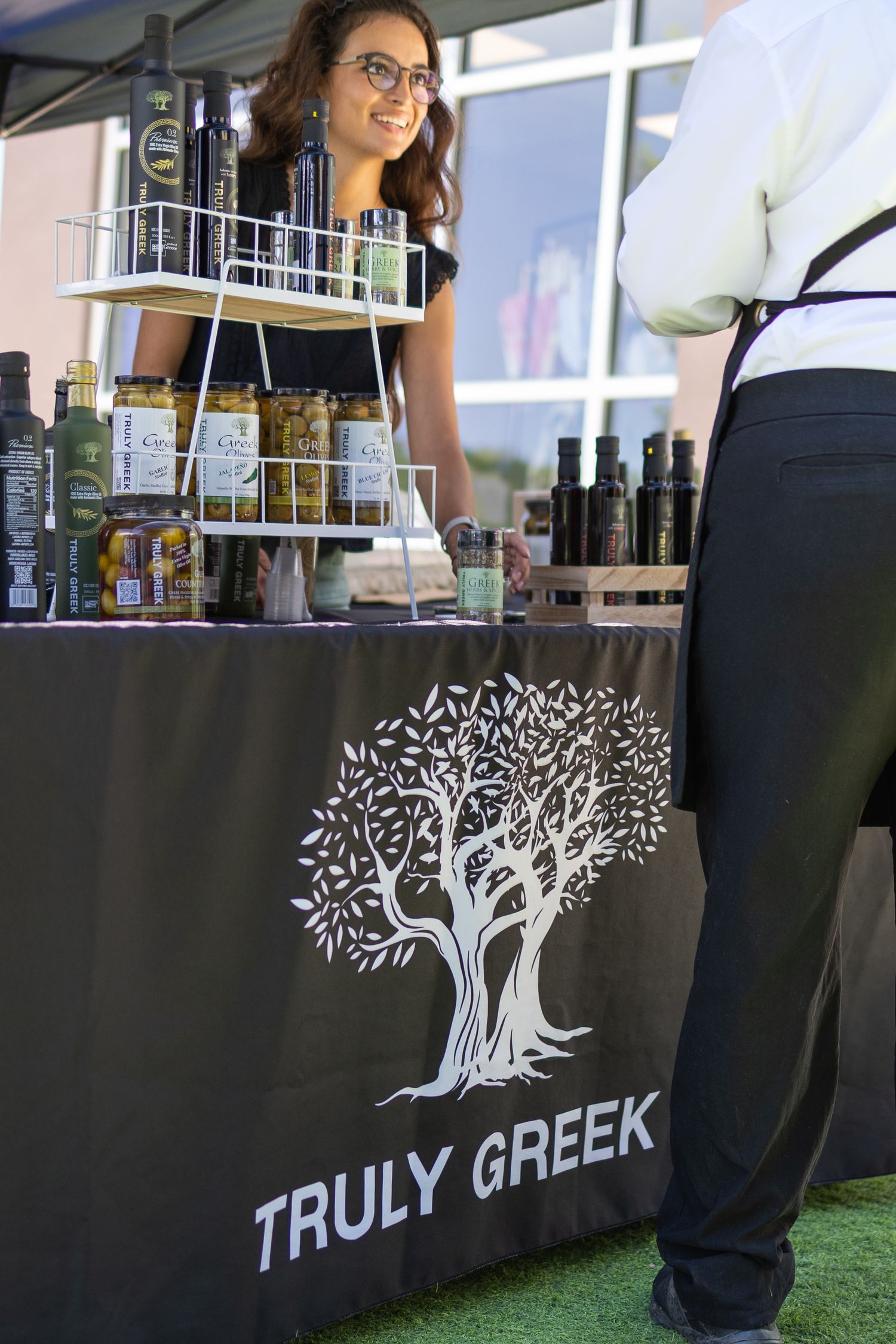 Truly Greek olive oil vendor booth with smiling woman showcasing Greek products at outdoor market.