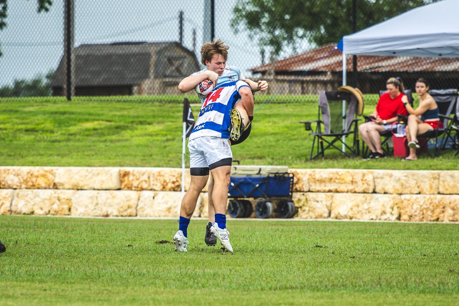 Rugby player in blue and white uniform kicking ball on green field during outdoor match with spectators watching.
