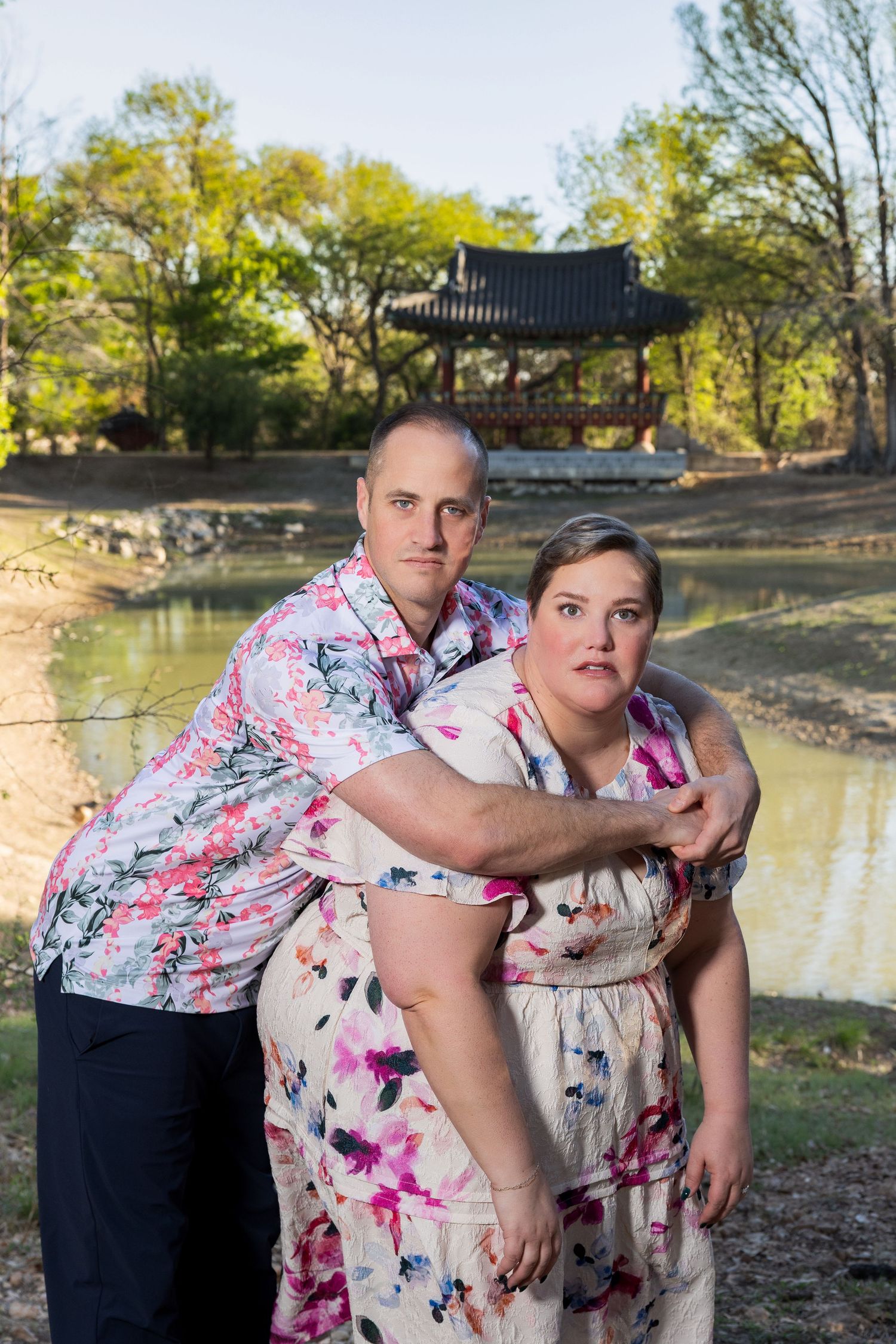 A man and woman wearing colorful floral patterned clothing stand together in a natural outdoor setting with trees and a body of water.