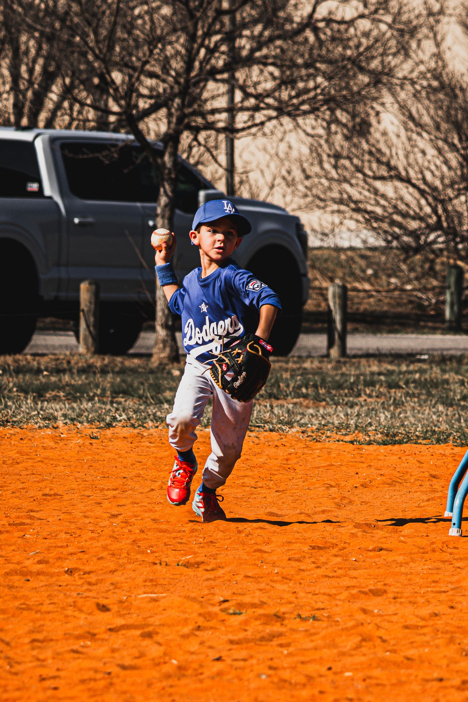 Young baseball player in Dodgers uniform throwing ball on red dirt infield during youth league game.