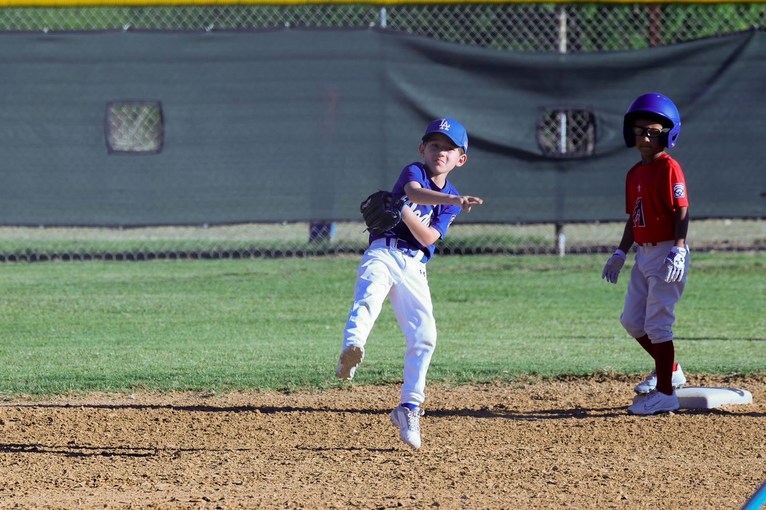 Youth baseball player throwing ball during game while opponent stands near second base on dirt infield.