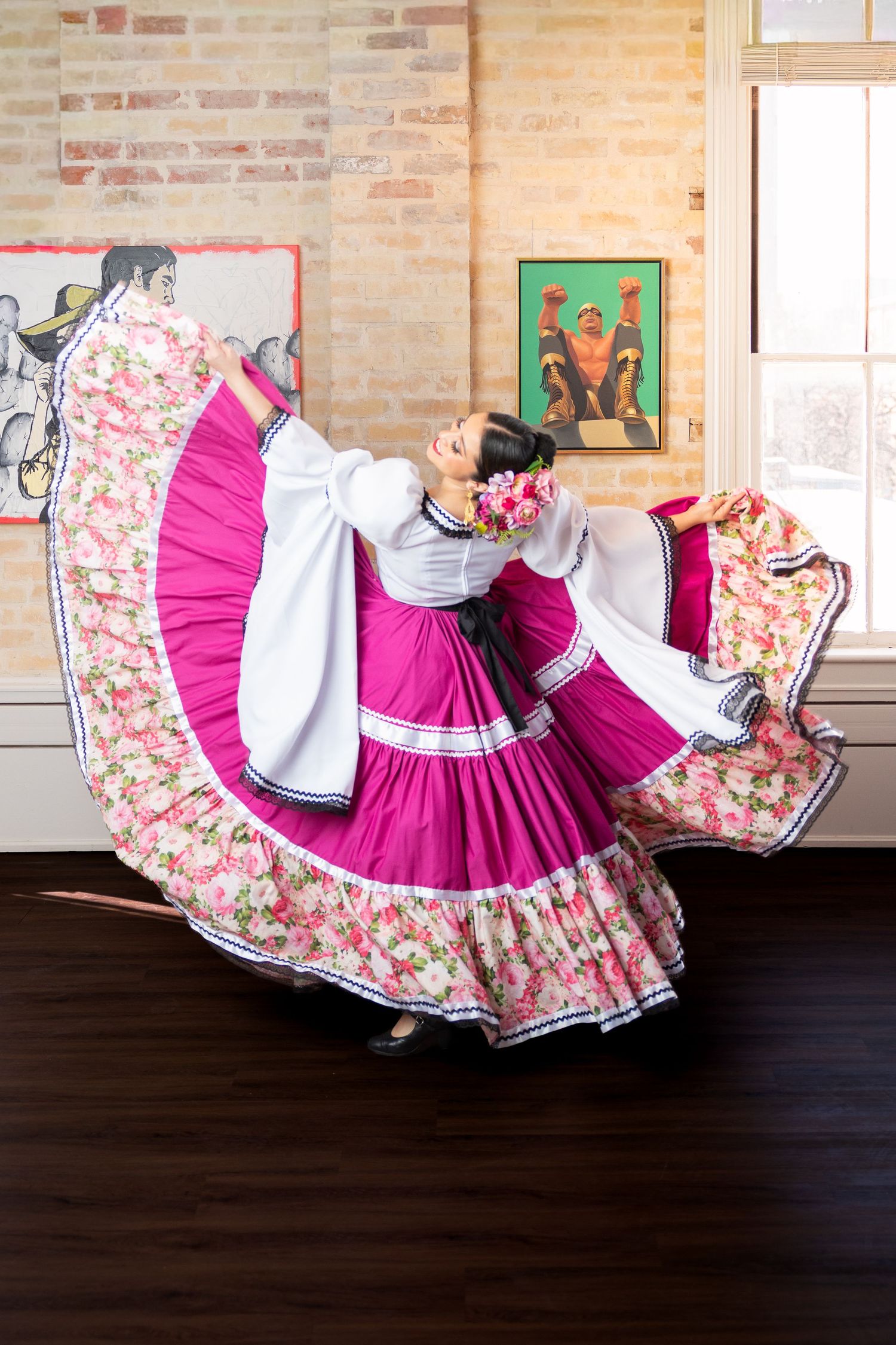 A woman in a vibrant pink and floral folkloric dress twirls gracefully in a dance studio with brick walls.