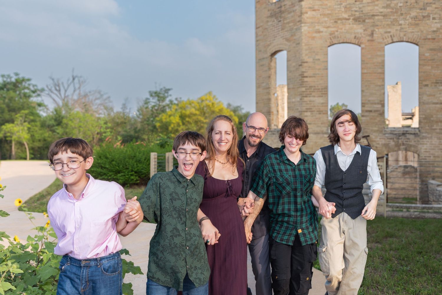 A smiling family of six poses outdoors near a historic stone ruins structure on a sunny day.
