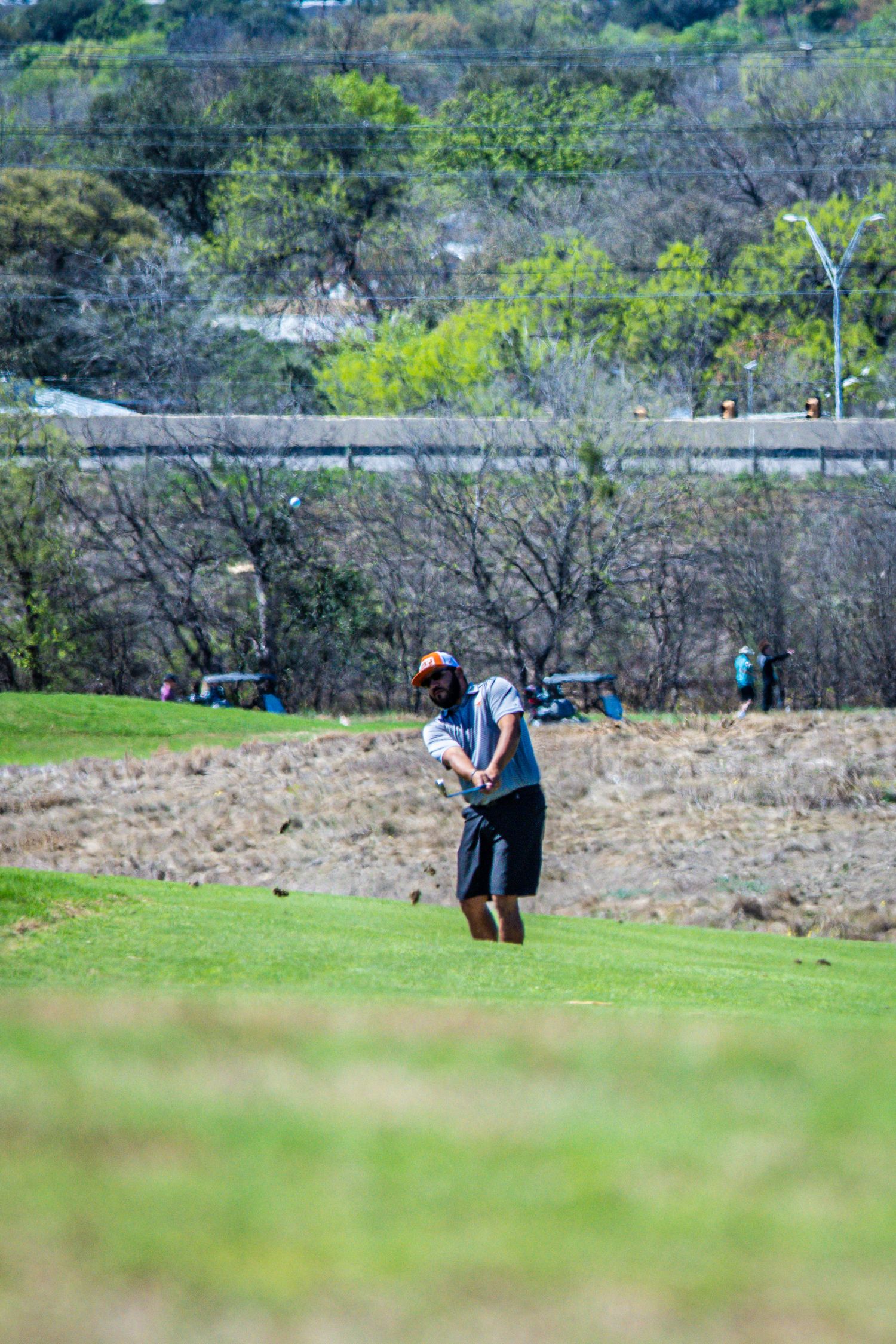 Golfer swinging a club on a lush green fairway with trees and hills in the background on a sunny day.
