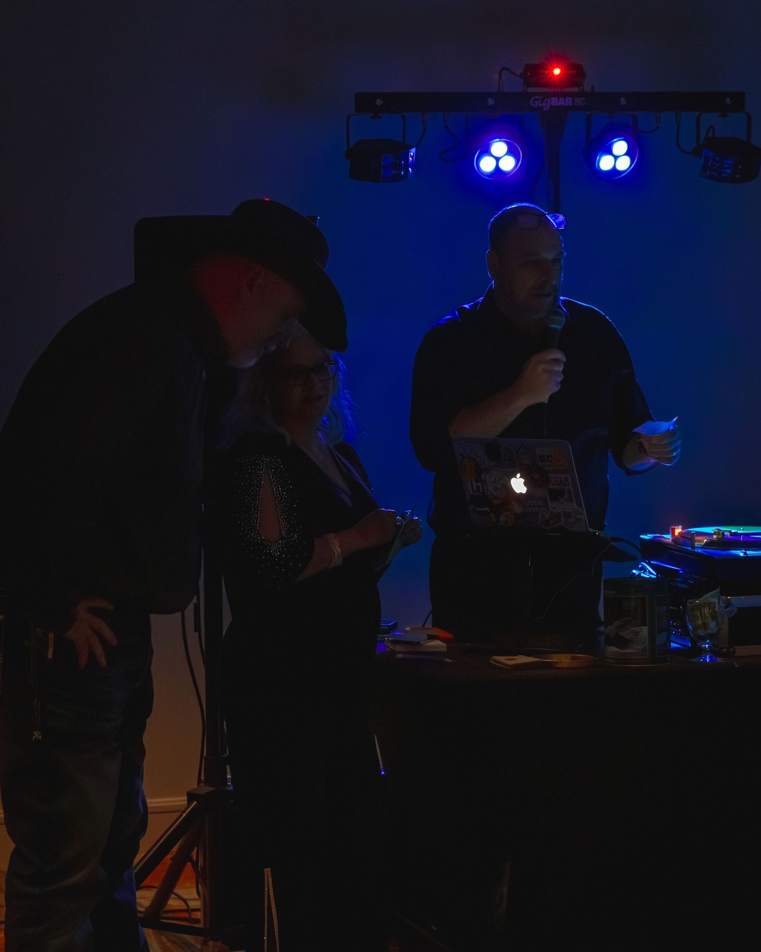 Three people gathered around a DJ table with a laptop and microphone under blue stage lights in a dark venue.