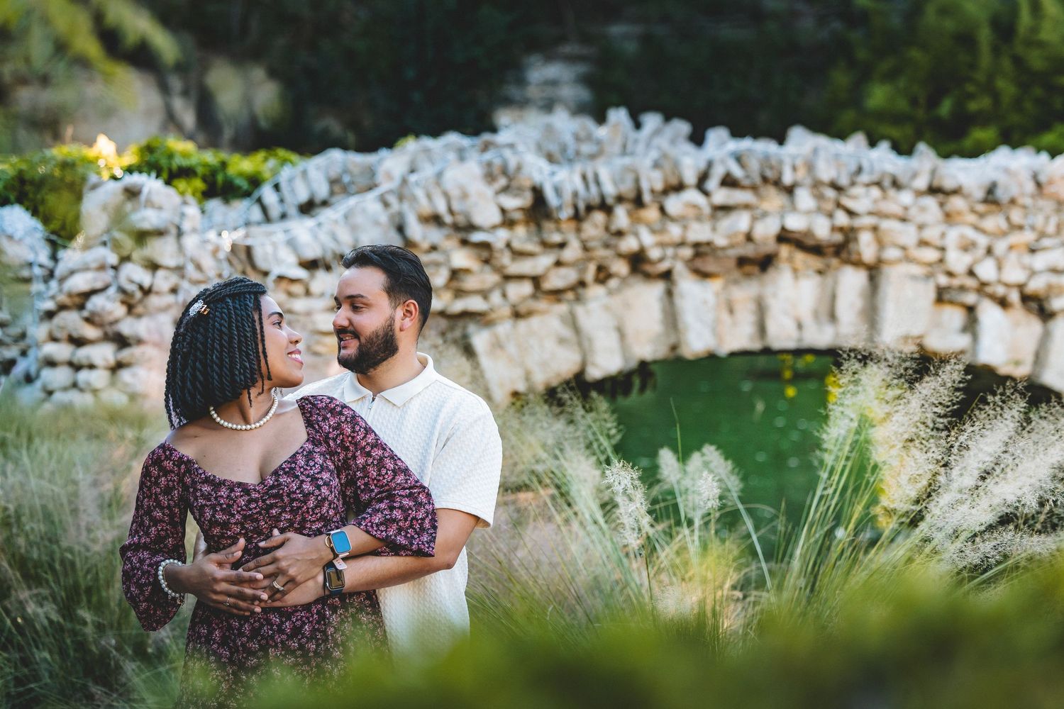 A couple shares an intimate moment in a rustic garden setting with stone walls and lush greenery in the background.