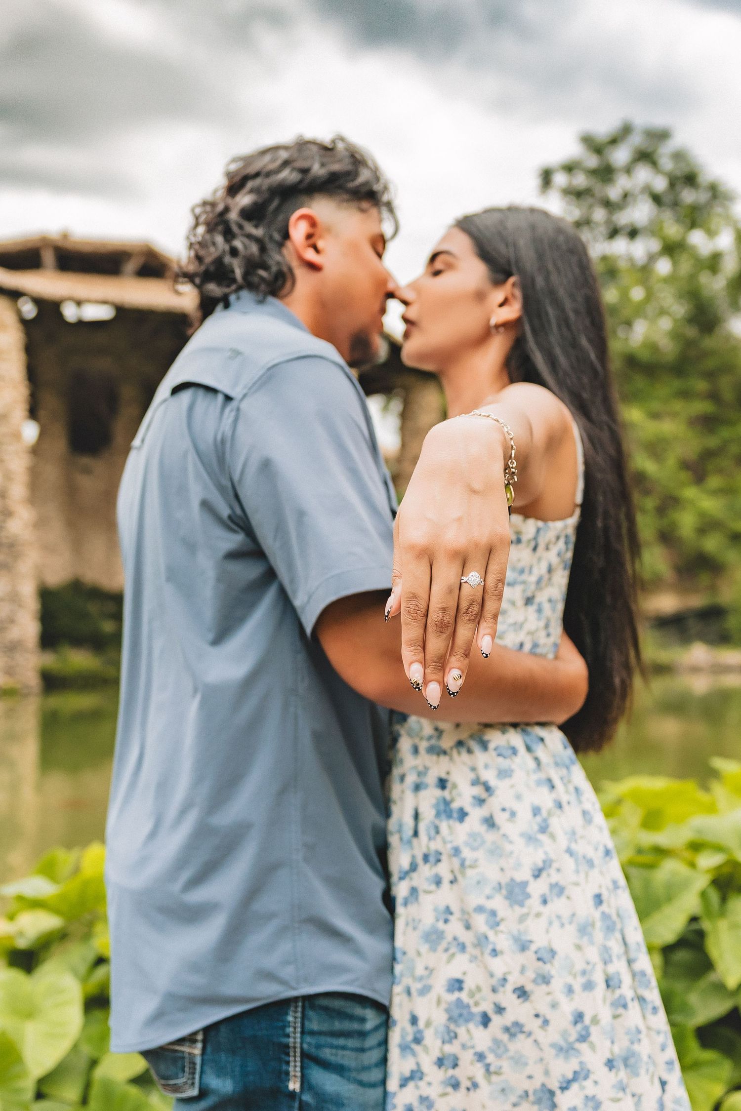 Couple kissing outdoors while woman shows off engagement ring, wearing floral dress near a pond.