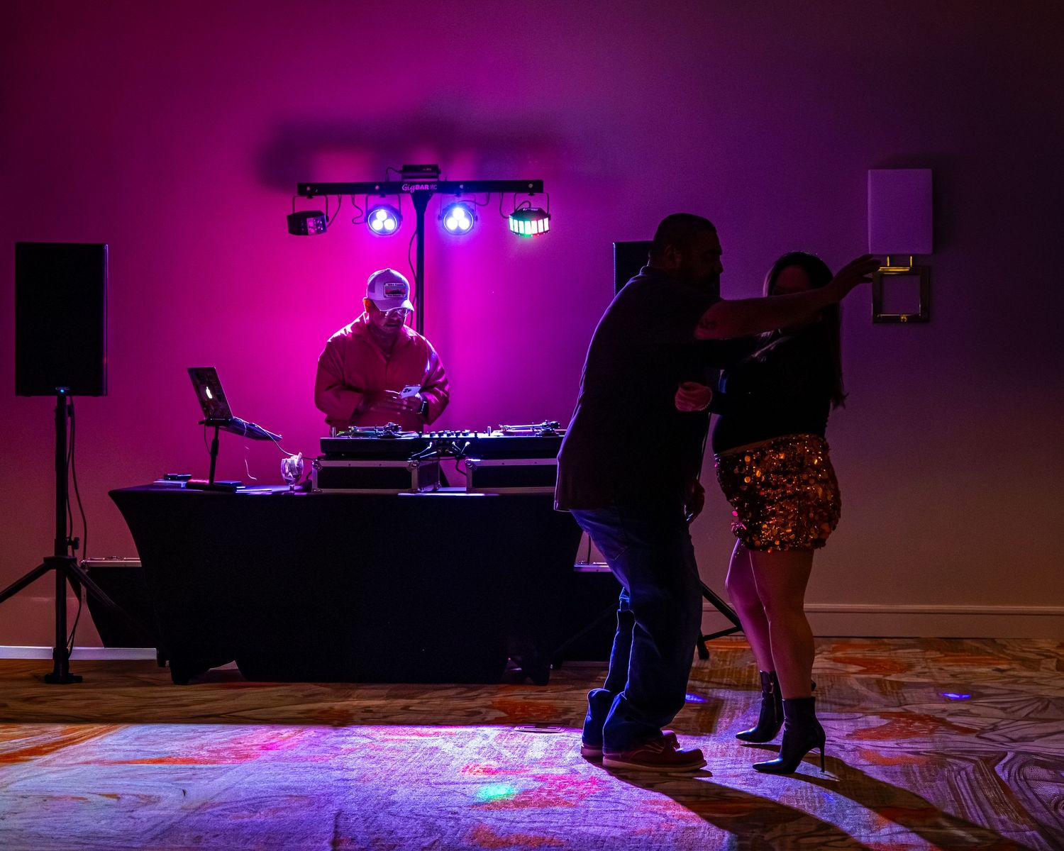 A DJ performs at a glowing pink-lit dance floor while two guests dance at an elegant indoor event.