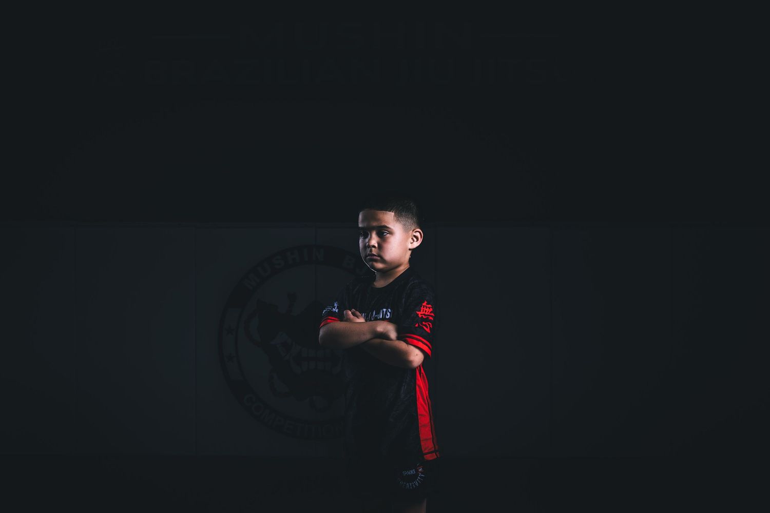 Young boy in red and black soccer jersey stands confidently with arms crossed against dark background.