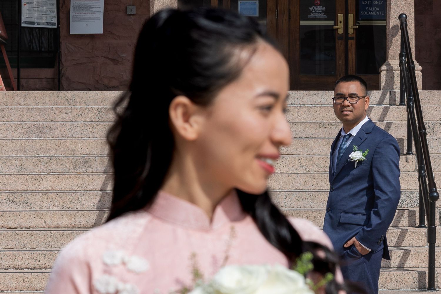 A wedding party poses on stone steps outside a brick building with the bride wearing a pink floral dress.