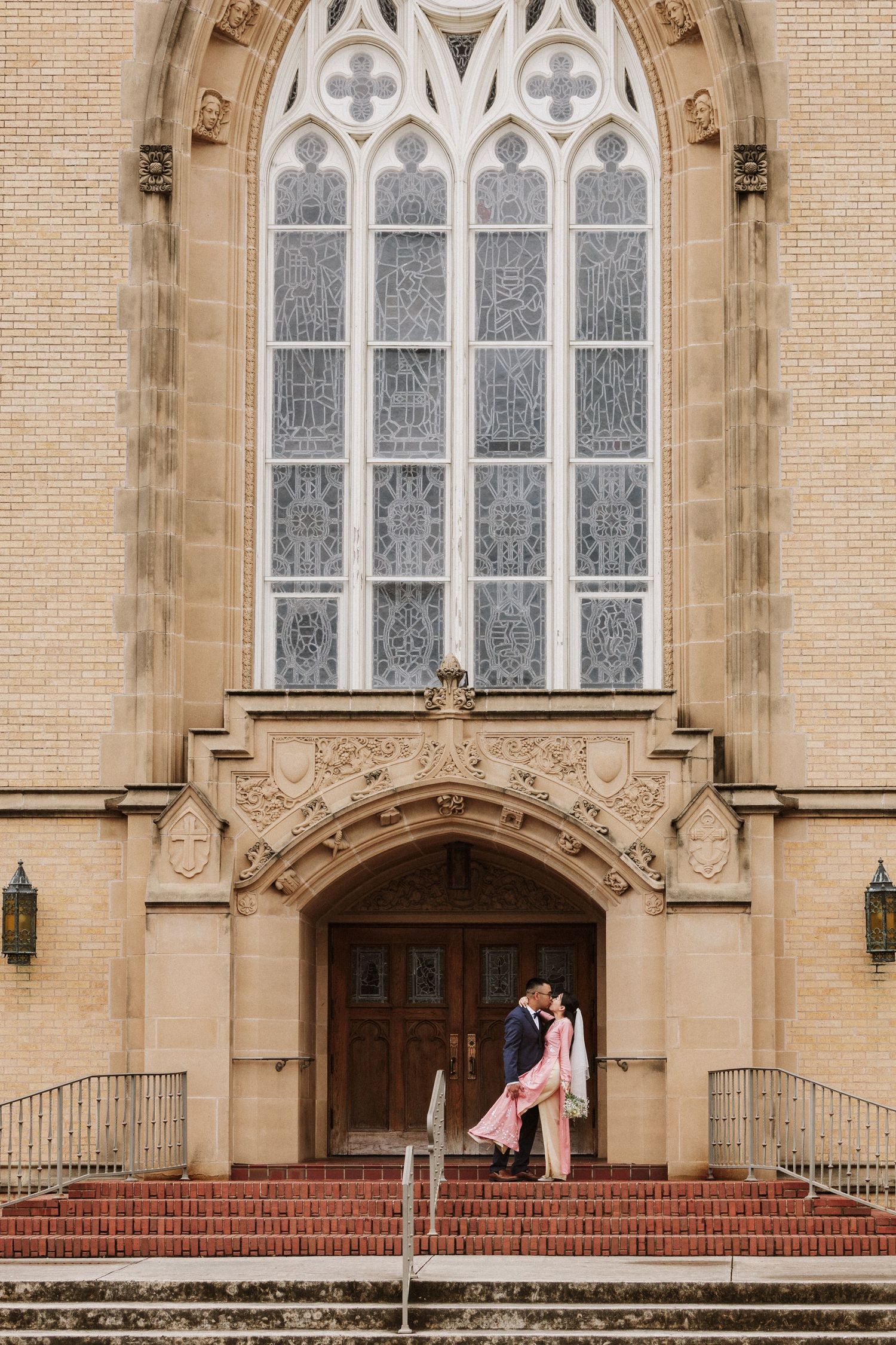A couple stands in front of a Gothic church entrance with ornate stone architecture and stained glass windows.