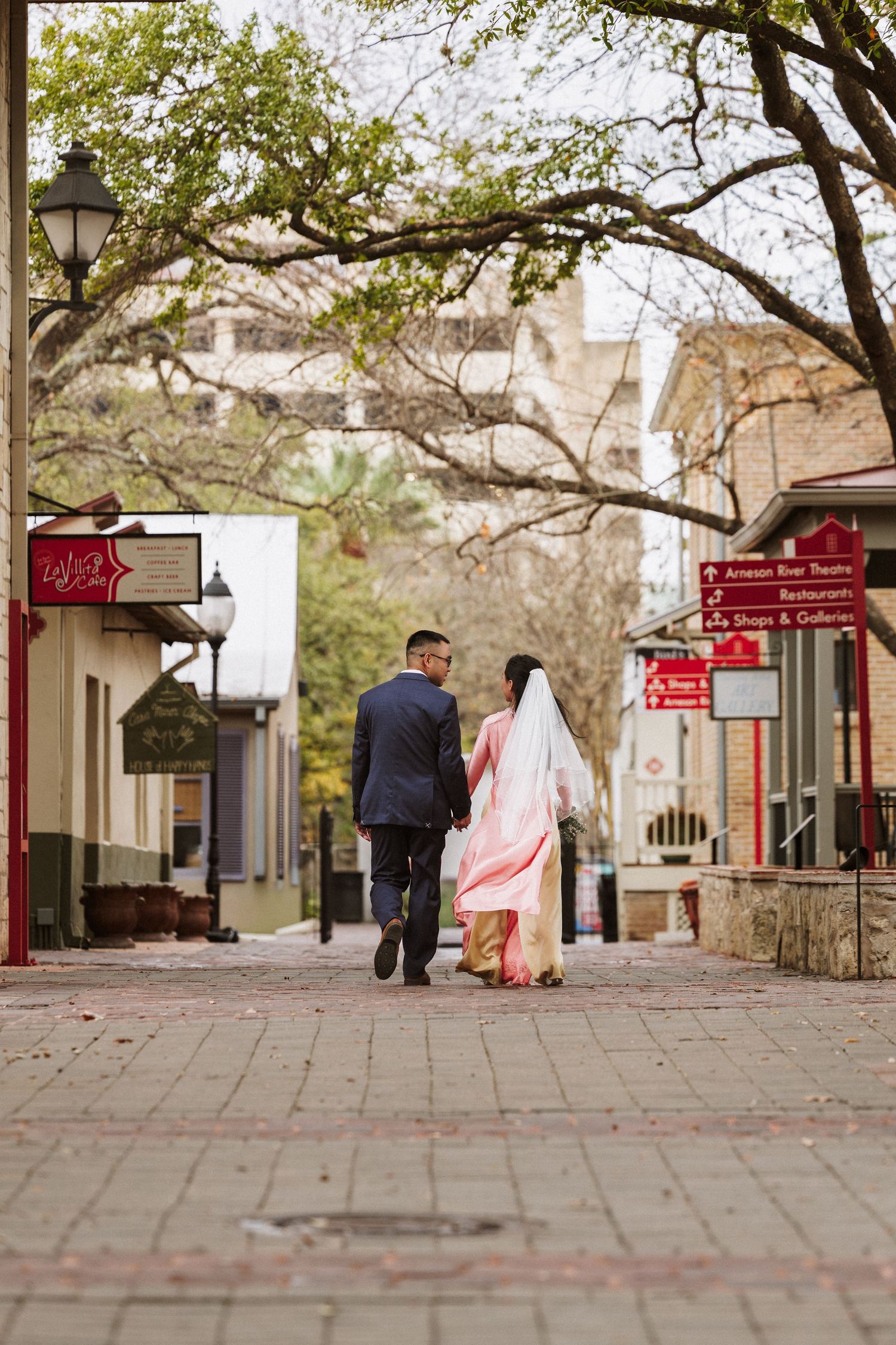 Couple in wedding attire walking hand in hand down a brick street in a historic town.