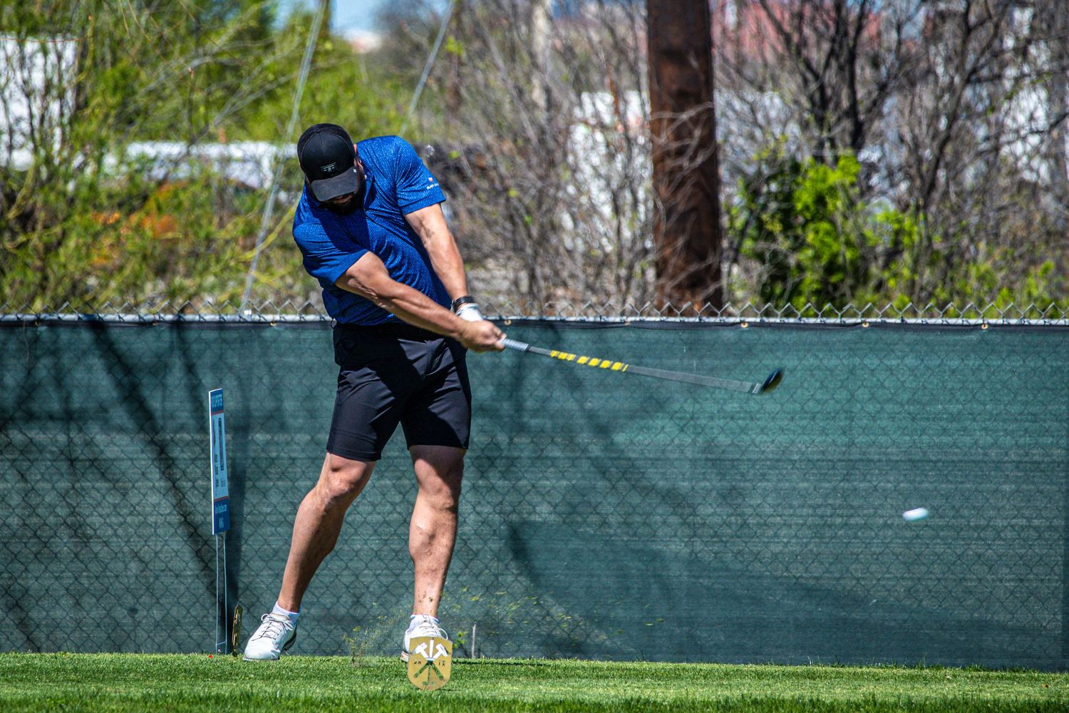 Golfer completing a powerful swing on a practice range with a chain-link fence backdrop.