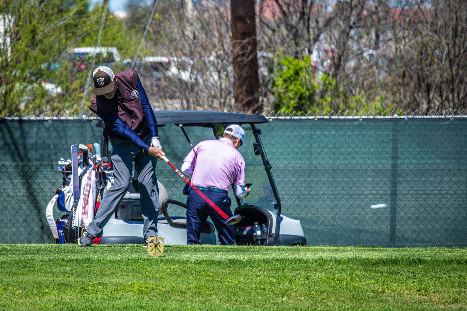 Two golfers near a golf cart and bag on a green course by a fence on a sunny day.