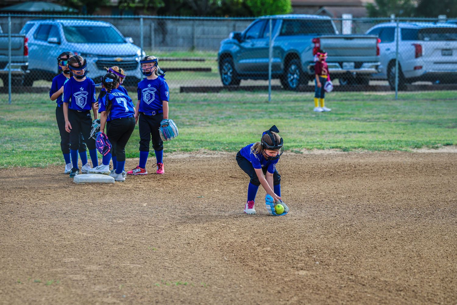 Young girls in blue uniforms playing softball on a dirt field, with one player fielding a ground ball near a base.