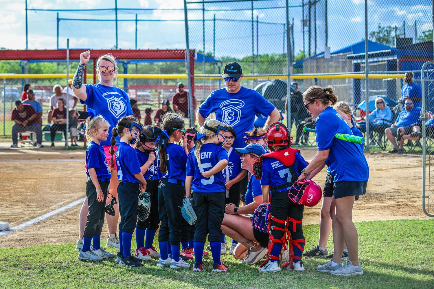 Youth softball team huddles with coaches in blue shirts on a sunny baseball field during a game.